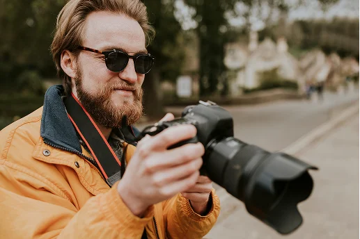 Een persoon met lang haar, die een bril draagt en een oranje jas aan heeft, houdt een DSLR-camera vast op een vervaagde straat, wat de focus op fotografie suggereert.
