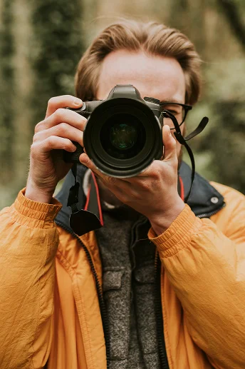 Een persoon in een gele jas houdt een camera voor hun gezicht en richt zich op het vastleggen van een afbeelding. De omgeving lijkt een natuurlijke buitenruimte te zijn.
