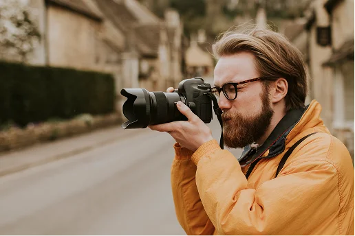 Een persoon met een baard die een bril draagt en een oranje jas aan heeft, is gefocust op het maken van een foto met een DSLR-camera. De achtergrond toont een pittoreske straat met stenen gebouwen, wat een vredige, landelijke setting suggereert.